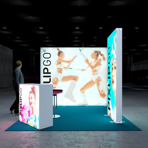 A colorful Pixlip GO exhibition booth featuring vibrant illuminated panels. The main backdrop shows two women joyfully playing with water and foam, while a side panel displays a portrait of a woman with foam bubbles in her hair. The front counter features a smiling woman holding a colorful toothbrush. A person observes the booth from a distance.